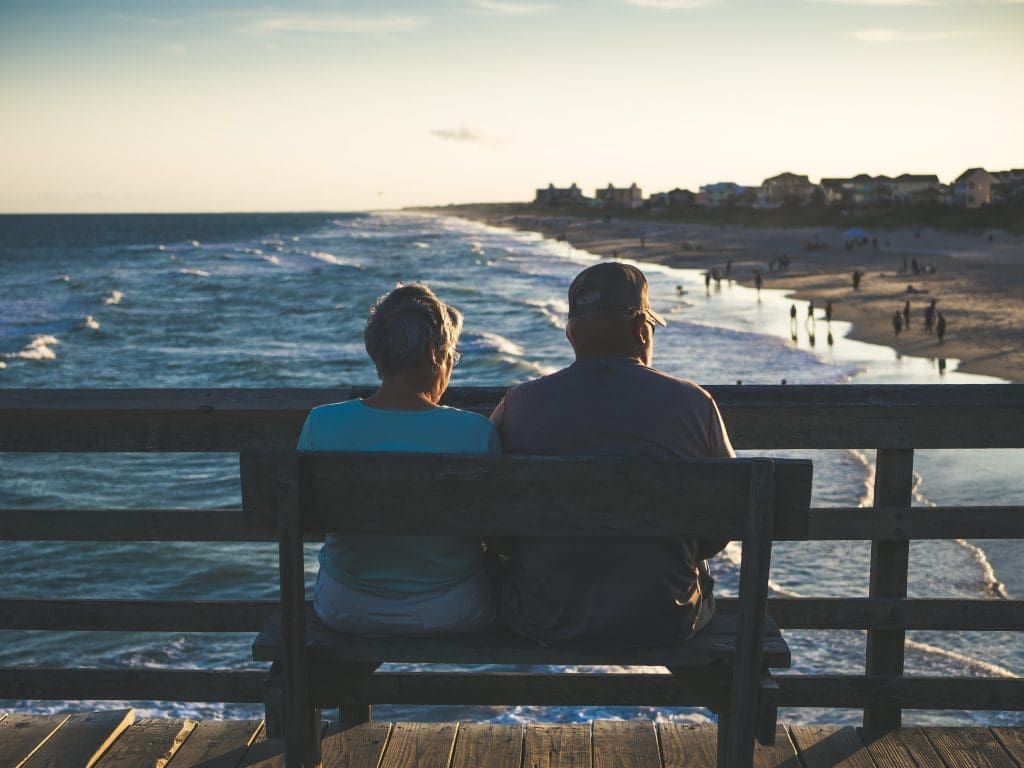 Planning for Retirement | Older Couple enjoying a view of the beach and sea from a bench up high