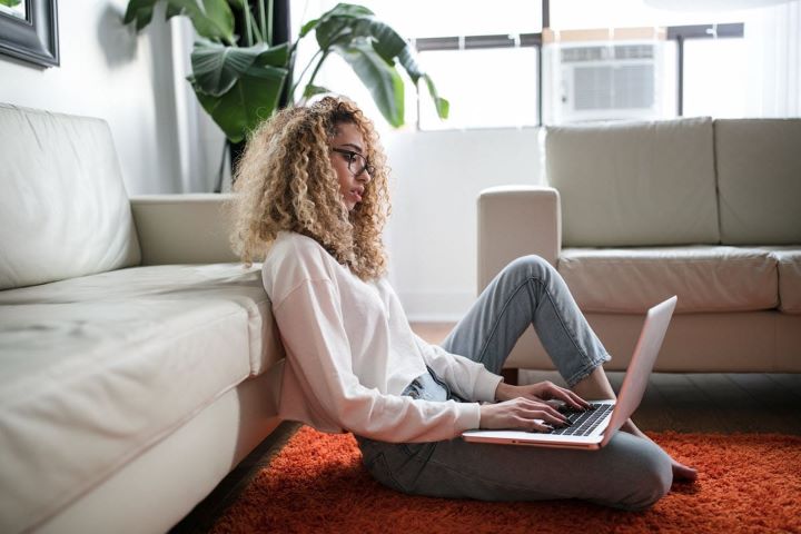 A lady sat on the floor leaning against a sofa working on her laptop