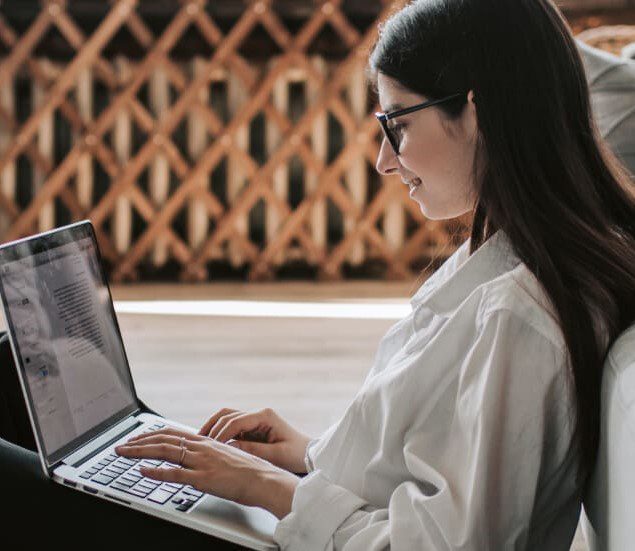 A lady sat on the floor leaning against a sofa working on her laptop