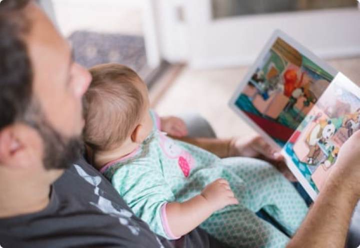 A baby sat on her dads lap while he reads a book to her